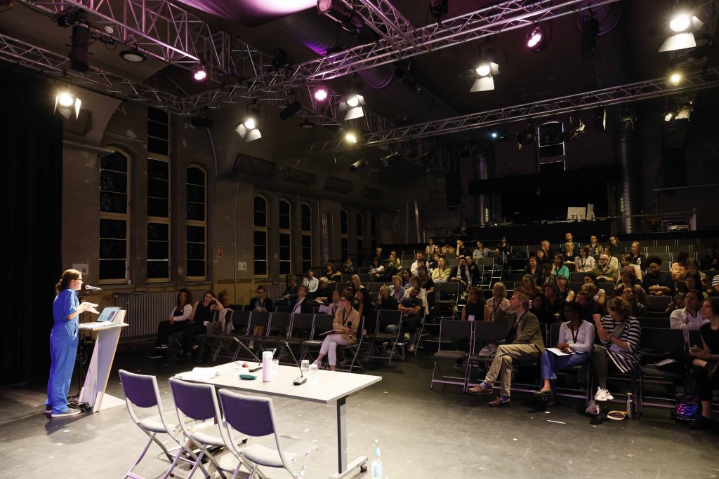 Blick ins Publikum des Theater Strahls, in dem die Connected Audience stattgefunden hat, mit einer vortragenden Person auf der Bühne.