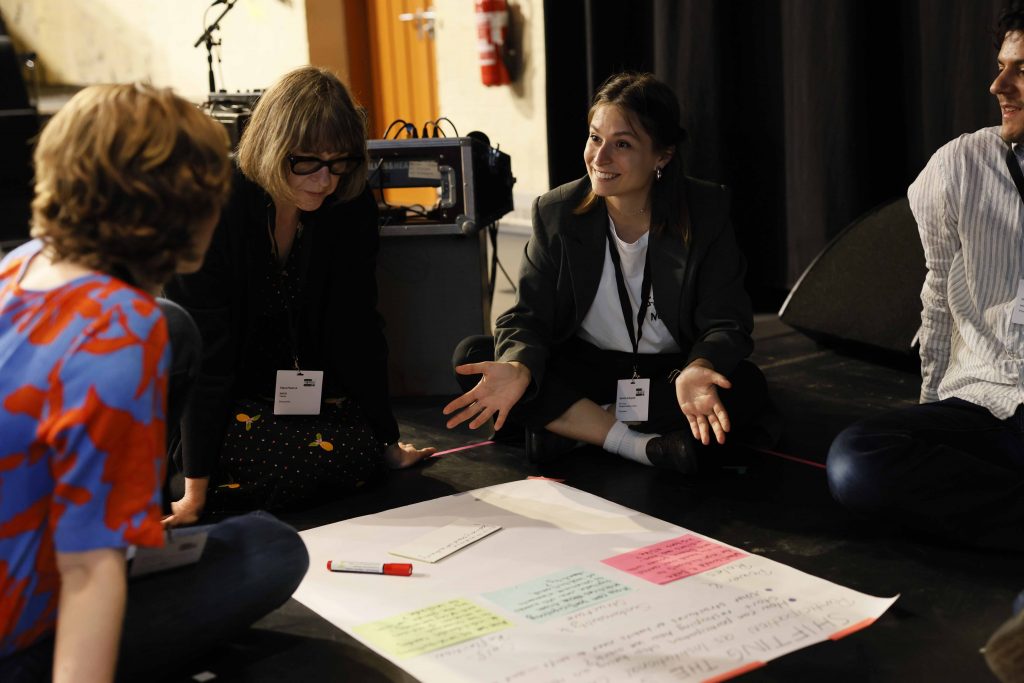 Gruppe von Menschen, die auf dem Boden sitzt und über ein Flipchart-Paper gebeugt diskutiert.
