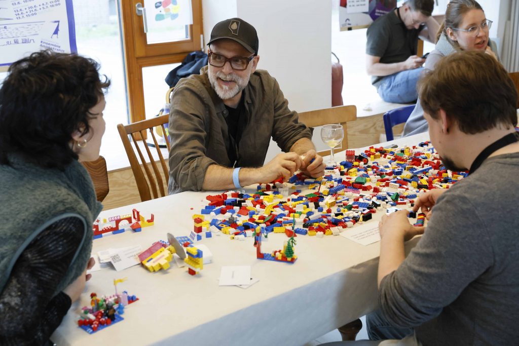 Ein Mann sitzt an einem Tisch vor Lego-Steinen und unterhält sich mit anderen Menschen.