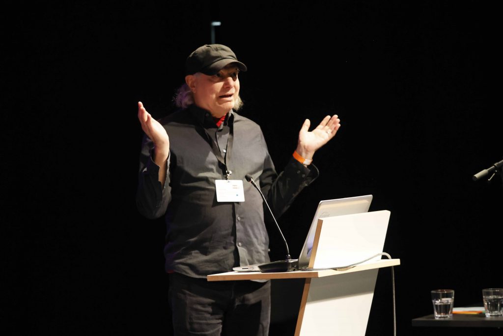 John H. Falk steht auf der Bühne und hält einen Vortrag.