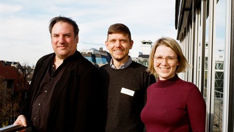 Drei Personen stehen auf einem Balkon und schauen in die Kamera.